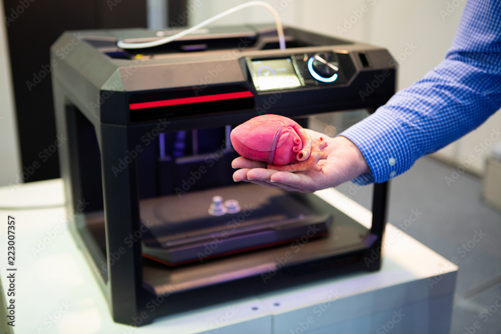 A man is holding a 3D-printed heart in front of a 3D printer